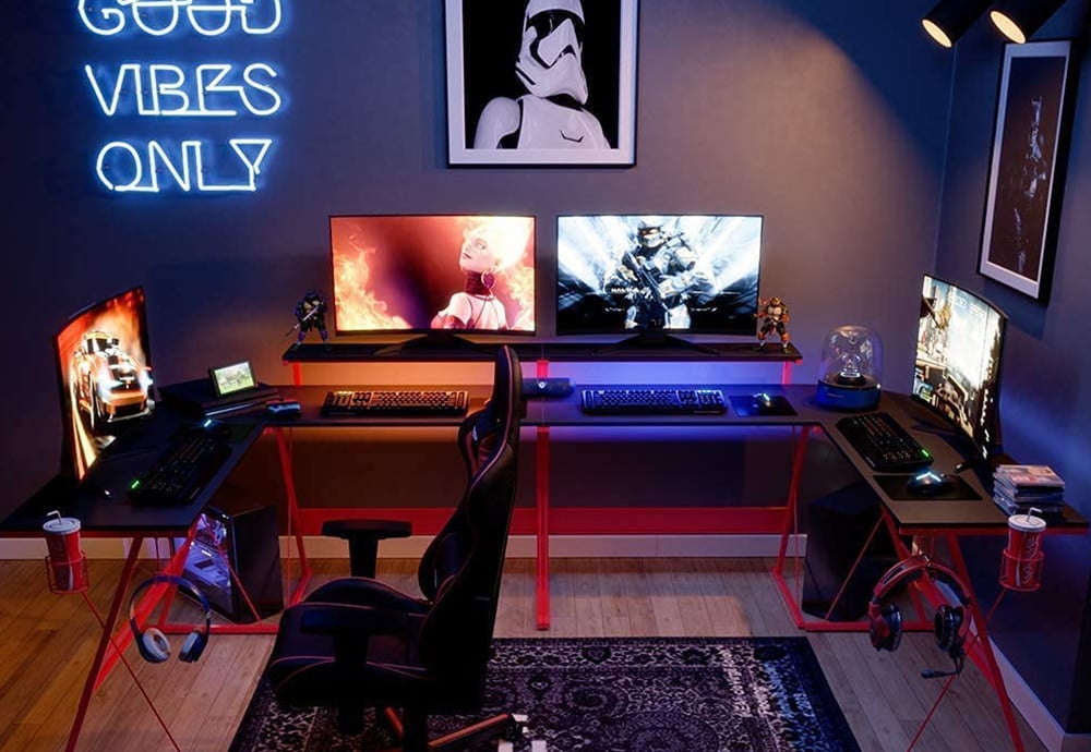 Two Bestier L-Shaped Gaming Desk RGB with LED lights, joined together to create a gaming station. On them there are 4 gaming monitors, with their corresponding gaming keyboards and mice. On the sides there are two hangers with headphones and two cup holders with soft drinks.