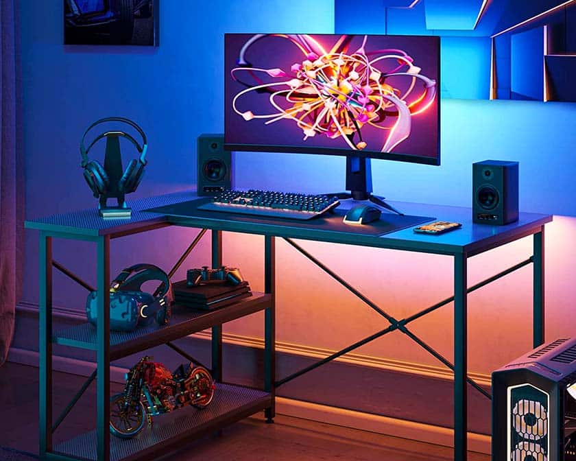 L-shaped Bestier gaming table with shelves, where various gaming devices are stored, such as VR glasses, a gamepad and a console. On the table are a headset, a gaming monitor and a pair of speakers, as well as a gaming keyboard and mouse.