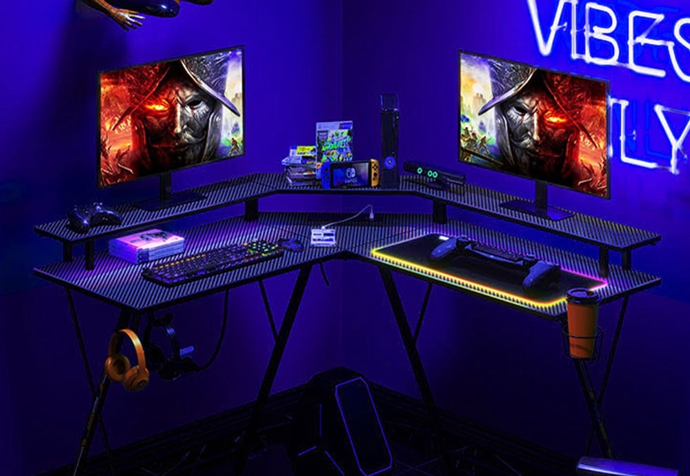 Seven Warrior L-shaped desk with LED strips, power sockets, headphone hook and drink holders. On top of the table, there are two monitors and their corresponding keyboards and game pads, as well as other gaming accessories.