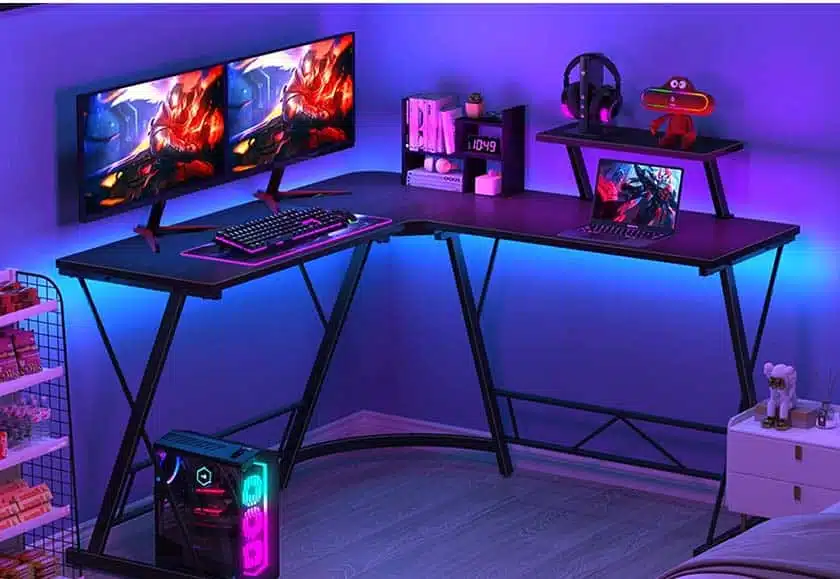 Mr IRONSTONE L-Shaped Desk, Computer Gaming Desk. On top, two monitors, a laptop, a headset and some accessories and decorative elements.