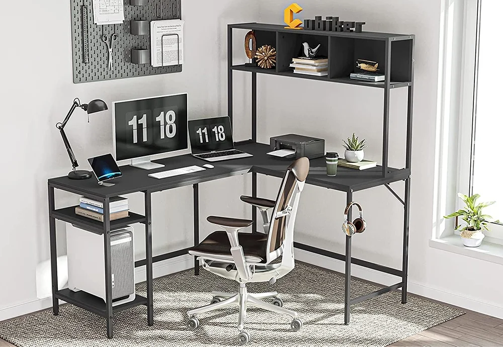 Escritorio en forma de L, de Cubiker, con un monitor, un portátil, una impresora y una tablet sobre la mesa. En el estante inferior hay una torre PC y en los superiores se muestran varios accesorios y libros. En el colgador hay unos auriculares de diadema.