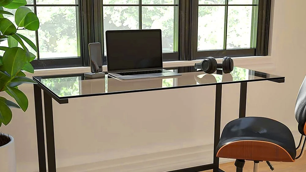 Escritorio con superficie de vidrio situado frente a una ventana, con un laptop, unos auriculares y un m&oacute;vil sobre &eacute;l.