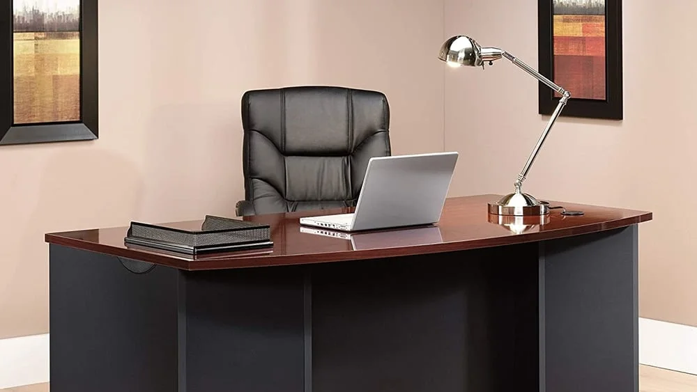 Escritorio ejecutivo con un ordenador port&aacute;til, una l&aacute;mpara de mesa y una bandeja para papeles sobre &eacute;l.